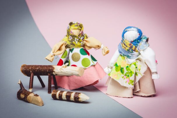 Kaks kanga‑nuku, mille pead katavad värvikirevad sallid, seismas roosa‑ ja hallil taustal; vasakul nukk kannab kollase täpilise mustriga salli ja roosat seelikut, paremal nukk on sinise salliga ja rohelise lillemustriga kleidiga; ees on pisikesed puidust kujukesed.