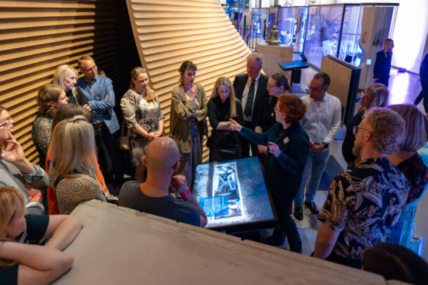 Rühm täiskasvanuid seisab ringis näitusesaalis; keskmes on suur puutetundlik ekraan, millel on sinakas pilt, ning üks esineja käega selgitab, teised kuulavad tähelepanelikult.