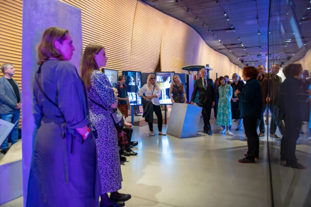 Galeriisaal, kus suur inimeste grupp seisab poolringis ning kuulab esinejat; keskmes on suur, nurgeline metallist plokk ning taustal on ekraanidel näidatud pilte ja sinakas-lilla valgus.