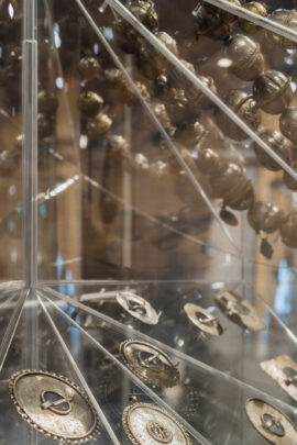 Klaasvitriinis on näha mitu ringikujulist metallmündi- või medaljoniplaatide; need on asetatud dekoratiivsetele aluspindadele ja vitriin peegeldab valgust, taustal on näha teisi eksponaate ja metallist üksikasju.