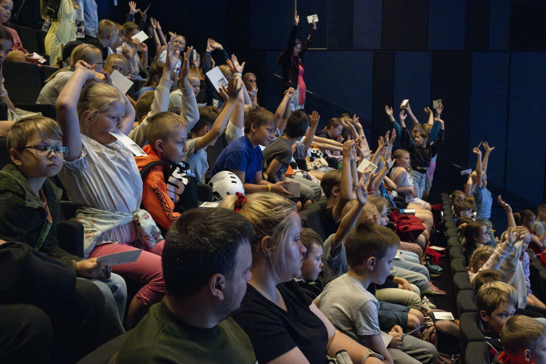Auditooriumis istub palju lapsi ja mõned täiskasvanud; suur hulk lapsi tõstab käsi.