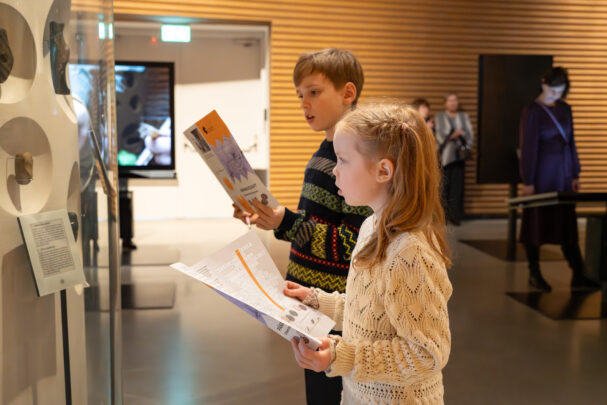 Kaks last, poiss ja tüdruk, seisavad näitusruumis klaasist vitriinide ees ja uurivad eksponaate, samal ajal hoides käes brošüüre.